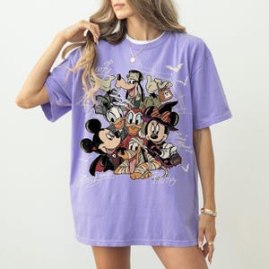 Może przedstawiać: Jasnofioletowy t-shirt z nadrukiem graficznym przedstawiającym postacie Disneya w kostiumach na Halloween, w tym Myszkę Miki jako wampira, Myszkę Minnie jako czarownicę, Goofy'ego jako Frankensteina i Pluto jako mumię, z białymi liniami.