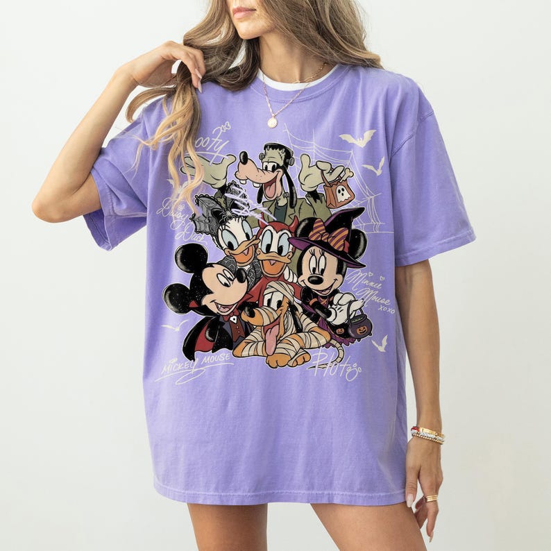 Może przedstawiać: Jasnofioletowy t-shirt z nadrukiem graficznym przedstawiającym postacie Disneya w kostiumach na Halloween, w tym Myszkę Miki jako wampira, Myszkę Minnie jako czarownicę, Goofy'ego jako Frankensteina i Pluto jako mumię, z białymi liniami.