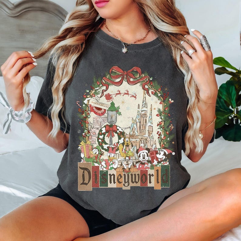 May include: Dark gray t-shirt featuring a colorful Disney World holiday design. The graphic includes a castle, characters, and the text "Disneyworld". The design is framed by a wreath with a red bow and Christmas decorations.