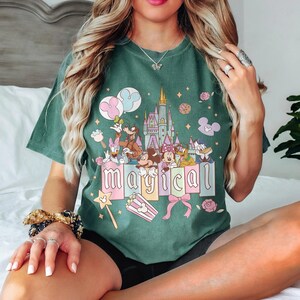 May include: Green t-shirt featuring a colorful graphic with Disney characters, a castle, and the word "magical." The design includes Mickey and Minnie Mouse, Donald Duck, and Goofy. Pink and blue balloons, popcorn, and a magic wand are also featured.