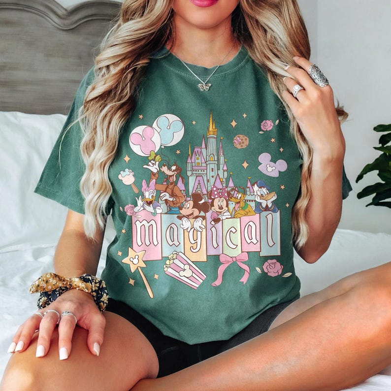 May include: Green t-shirt featuring a colorful graphic with Disney characters, a castle, and the word "magical." The design includes Mickey and Minnie Mouse, Donald Duck, and Goofy. Pink and blue balloons, popcorn, and a magic wand are also featured.