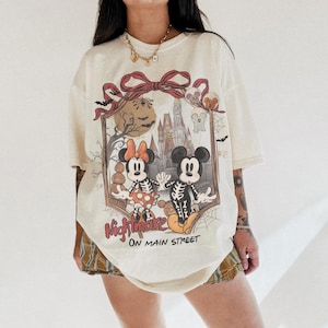 May include: Cream-colored t-shirt featuring a Halloween-themed graphic of Mickey and Minnie Mouse as skeletons in front of a haunted castle. The text "Nightmare on Main Street" is below the graphic. The shirt has short sleeves and a relaxed fit.