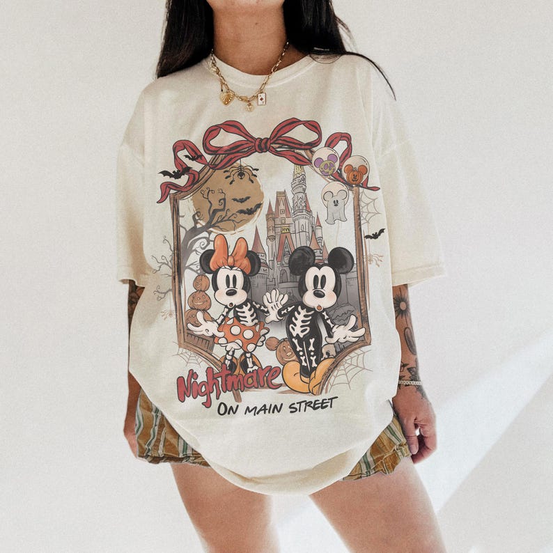 May include: Cream-colored t-shirt featuring a Halloween-themed graphic of Mickey and Minnie Mouse as skeletons in front of a haunted castle. The text "Nightmare on Main Street" is below the graphic. The shirt has short sleeves and a relaxed fit.
