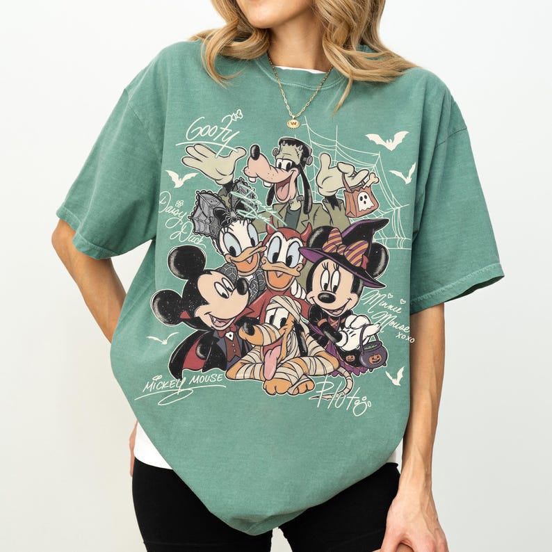 Może przedstawiać: Szałwiowo-zielony t-shirt z kr&oacute;tkim rękawem z nadrukiem graficznym przedstawiającym postacie Disneya w kostiumach na Halloween, w tym Myszkę Miki, Myszkę Minnie, Goofy'ego, Kaczora Donalda, Daisy i Pluto, z pajęczynami, nietoperzami i napisami.