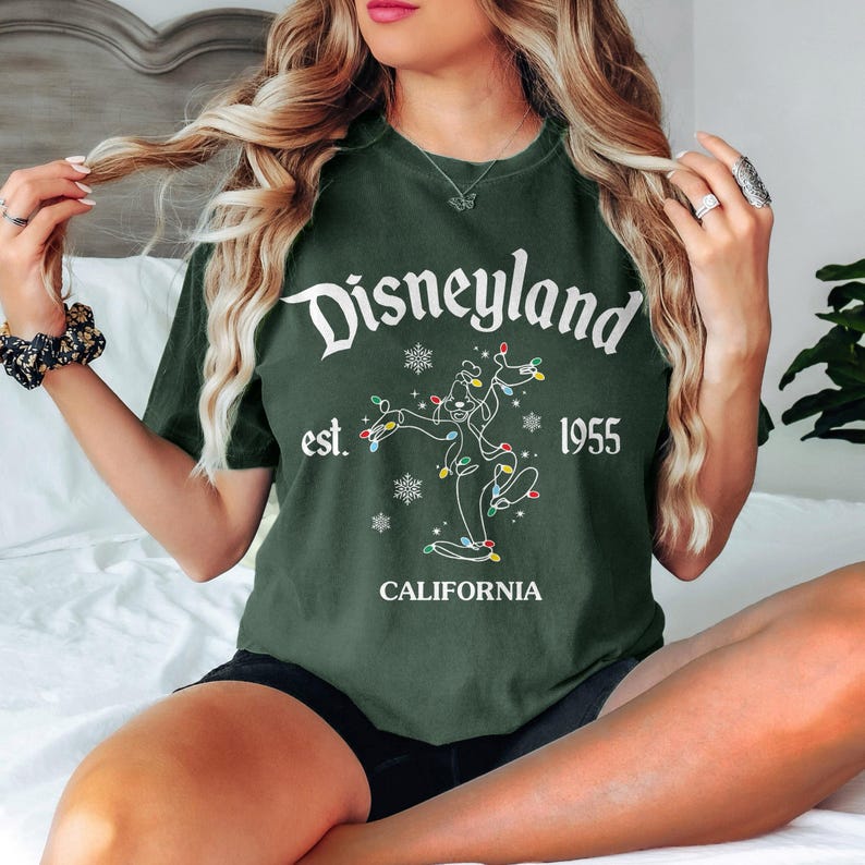May include: Dark green t-shirt with the Disneyland logo in white script. The shirt features a graphic of a character with Christmas lights and the text "est. 1955 CALIFORNIA". The shirt is a relaxed fit.