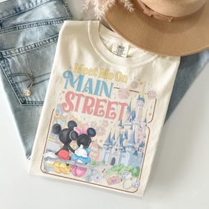 Op de afbeelding: Cr&egrave;mekleurig T-shirt met de tekst "Meet Me On Main Street" in geel, blauw en roze. De afbeelding toont Mickey en Minnie Mouse, een kasteel en vuurwerk. Een bruine hoed en een blauwe jeans staan op de achtergrond.
