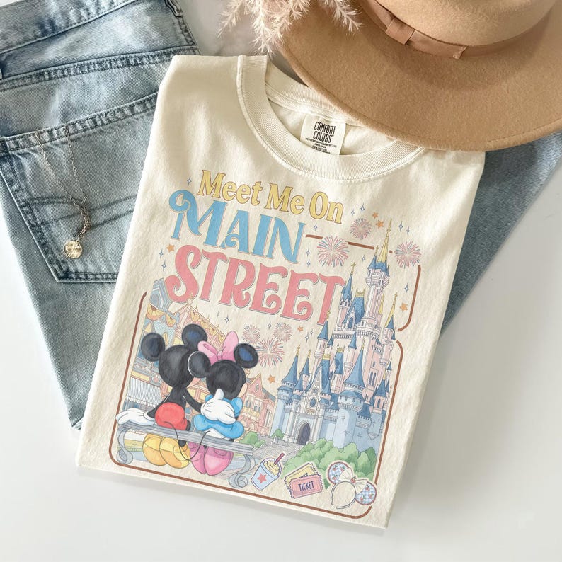 Op de afbeelding: Cr&egrave;mekleurig T-shirt met de tekst "Meet Me On Main Street" in geel, blauw en roze. De afbeelding toont Mickey en Minnie Mouse, een kasteel en vuurwerk. Een bruine hoed en een blauwe jeans staan op de achtergrond.