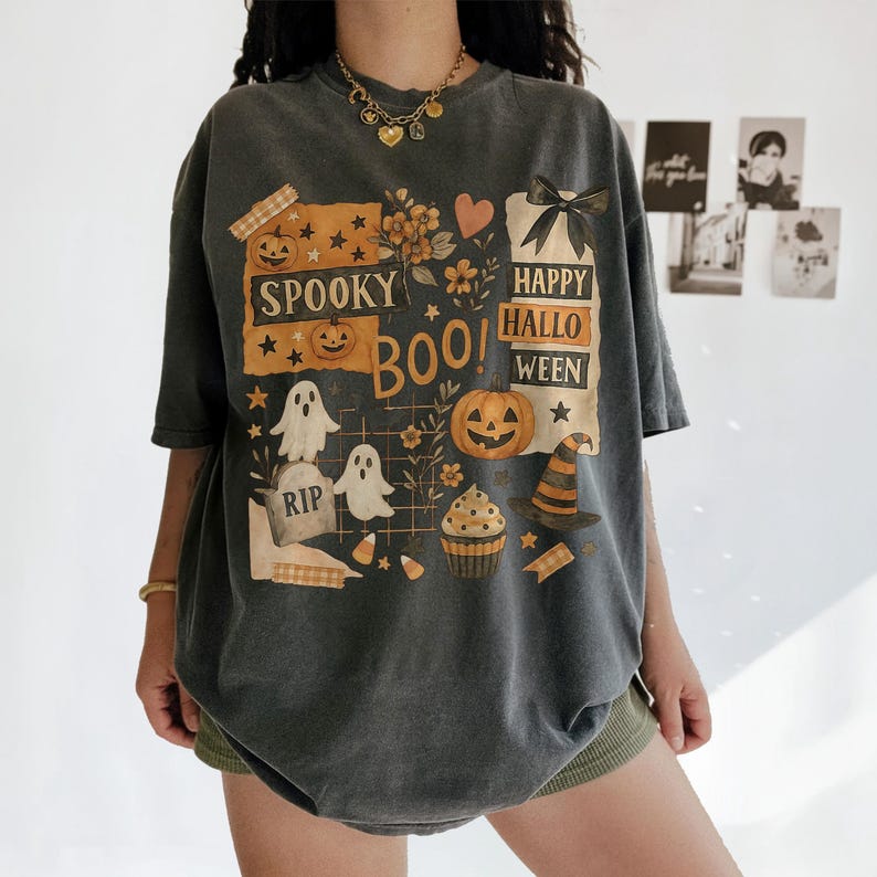 K&ouml;nnte beinhalten: Dunkelgraues Halloween-T-Shirt mit einem grafischen Design mit K&uuml;rbissen, Geistern, einem Hexenhut und den Worten "Spooky Boo! Happy Halloween". Das Design umfasst Sterne, Blumen und einen Grabstein.
