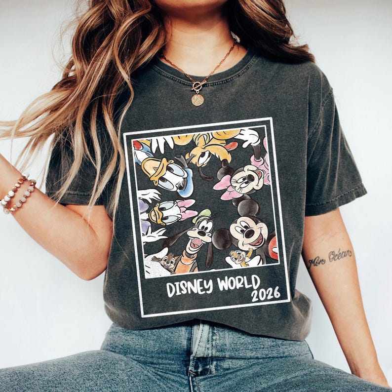 Puede incluir: Camiseta gris oscuro con un gr&aacute;fico enmarcado de personajes de Disney, incluyendo Mickey Mouse, Minnie Mouse, Donald Duck y Goofy. El texto "DISNEY WORLD 2026" est&aacute; impreso en blanco debajo.