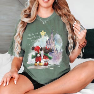 Pu&ograve; includere: T-shirt verde salvia con una grafica festiva di Topolino e Minnie Mouse in abiti natalizi, davanti a un castello. Il testo "It's the most magical time of the year" &egrave; stampato sopra i personaggi.