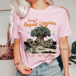 May include: A light pink t-shirt featuring the text "Disney's Animal Kingdom" in orange. The shirt has a graphic of Mickey Mouse in safari attire in front of the Tree of Life. The text "Wild about Disney" is below the graphic.