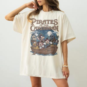 K&ouml;nnte beinhalten: Cremefarbenes T-Shirt mit einem Aufdruck eines Bootes mit Zeichentrickfiguren, darunter Micky Maus, Goofy und ein Totenkopf, mit dem Text "Pirates of the Caribbean" in Braun und Blau. Das Shirt hat kurze &Auml;rmel und eine lockere Passform.