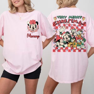 May include: Pale pink t-shirts with festive graphics. One shirt displays Minnie Mouse with the word "Mommy". The other features a "Very Merry Christmas" design with Mickey, Minnie, Goofy, Donald, and Pluto. Both have short sleeves.