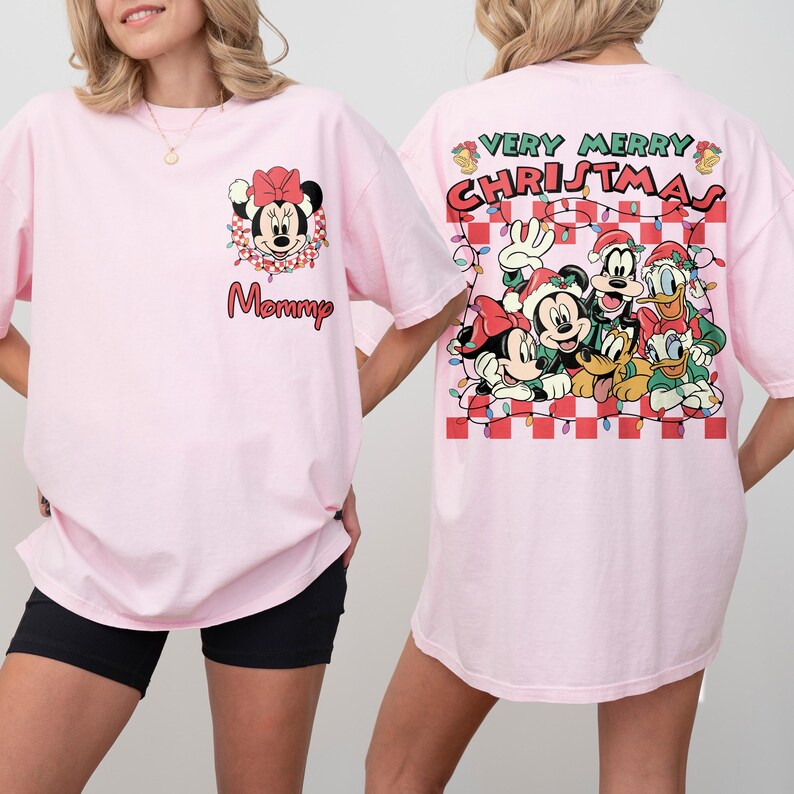 May include: Pale pink t-shirts with festive graphics. One shirt displays Minnie Mouse with the word "Mommy". The other features a "Very Merry Christmas" design with Mickey, Minnie, Goofy, Donald, and Pluto. Both have short sleeves.