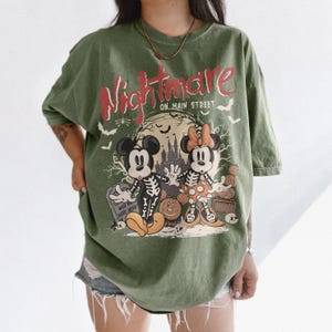 May include: Olive green t-shirt featuring a Halloween-themed graphic. The design includes Mickey and Minnie Mouse dressed as skeletons, a spooky castle, and the text "Nightmare on Main Street" in red and white. Bats and a tombstone complete the design.