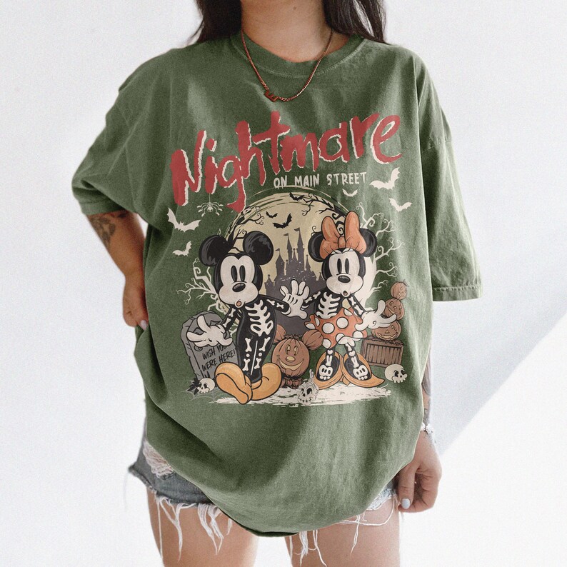 May include: Olive green t-shirt featuring a Halloween-themed graphic. The design includes Mickey and Minnie Mouse dressed as skeletons, a spooky castle, and the text "Nightmare on Main Street" in red and white. Bats and a tombstone complete the design.