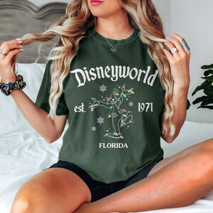 May include: Dark green t-shirt featuring the text "Disneyworld" in white script, with "est. 1971" and "Florida". A line drawing of a character is decorated with colourful lights and snowflakes.