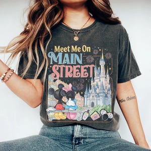 Op de afbeelding: Donkergrijs t-shirt met de tekst "Meet Me On Main Street" in kleurrijke letters. De afbeelding toont Mickey en Minnie Mouse, een kasteel, vuurwerk en andere parkelementen. Het shirt heeft een vintage stijl.