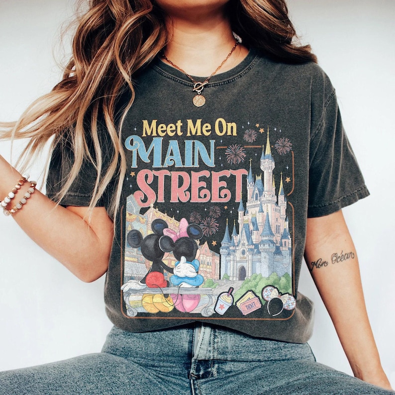 Op de afbeelding: Donkergrijs t-shirt met de tekst "Meet Me On Main Street" in kleurrijke letters. De afbeelding toont Mickey en Minnie Mouse, een kasteel, vuurwerk en andere parkelementen. Het shirt heeft een vintage stijl.