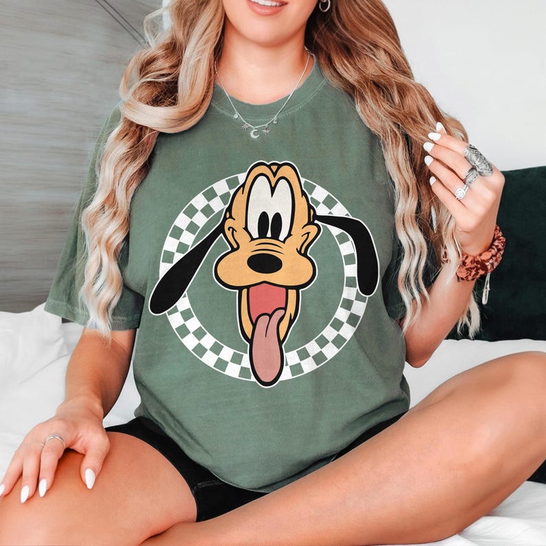 May include: Sage green t-shirt with a cartoon Pluto graphic. The graphic shows Pluto's face with his tongue sticking out, set against a white and black checkered circle. The shirt is a casual style.