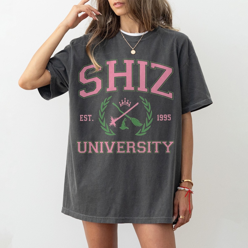 Pu&ograve; includere: T-shirt grigio scuro con la scritta "SHIZ UNIVERSITY" in rosa e verde. La maglietta presenta un design in stile collegiale con uno stemma che include scope incrociate, una corona e l'anno 1995.