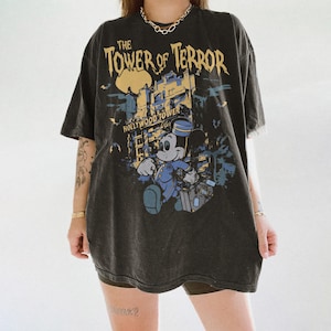 May include: Dark gray t-shirt featuring a graphic of Mickey Mouse in a bellhop uniform, with the text "The Tower of Terror" and "Hollywood Tower". The design includes a building and bats.
