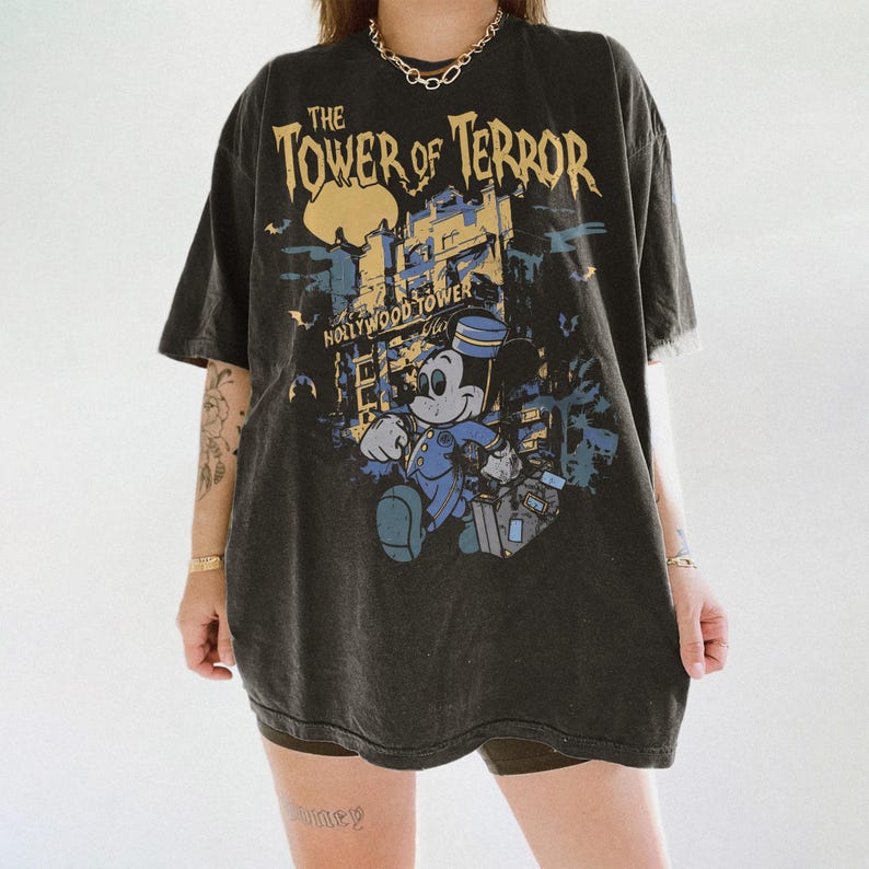 May include: Dark gray t-shirt featuring a graphic of Mickey Mouse in a bellhop uniform, with the text "The Tower of Terror" and "Hollywood Tower". The design includes a building and bats.