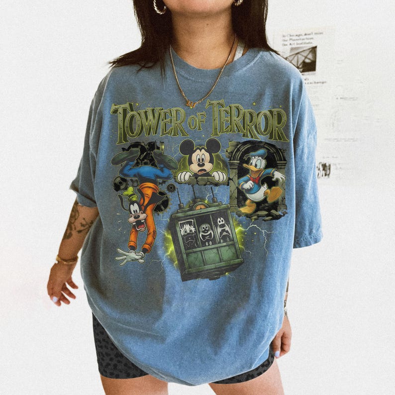 May include: A light blue short-sleeve t-shirt featuring a graphic print with the text Tower of Terror. The design includes illustrations of Goofy, Mickey Mouse, and Donald Duck surrounding a green elevator car with lightning accents.