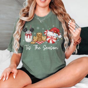 May include: Green t-shirt with a festive design featuring a milk jar with Mickey Mouse ears, cookies, and a candy cane snowman wearing a Santa hat. The text "Tis The Season" is printed below the graphic.