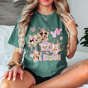 May include: Green t-shirt featuring Disney characters Mickey, Minnie, Daisy, Donald, and Pluto inside star-shaped frames. The word "magical" is below the characters. Pink and yellow accents and balloons complete the design.