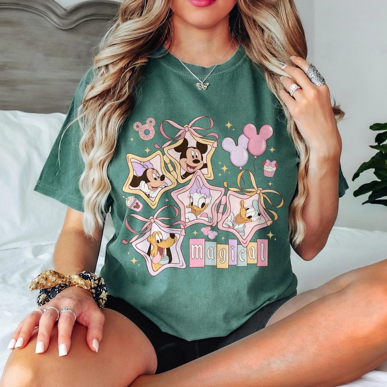 May include: Green t-shirt featuring Disney characters Mickey, Minnie, Daisy, Donald, and Pluto inside star-shaped frames. The word "magical" is below the characters. Pink and yellow accents and balloons complete the design.
