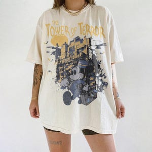 May include: Cream-colored t-shirt featuring a graphic of Mickey Mouse in a bellhop uniform, with the text "The Tower of Terror" and "Hollywood Tower Hotel". The design includes a stylized image of a building and bats.