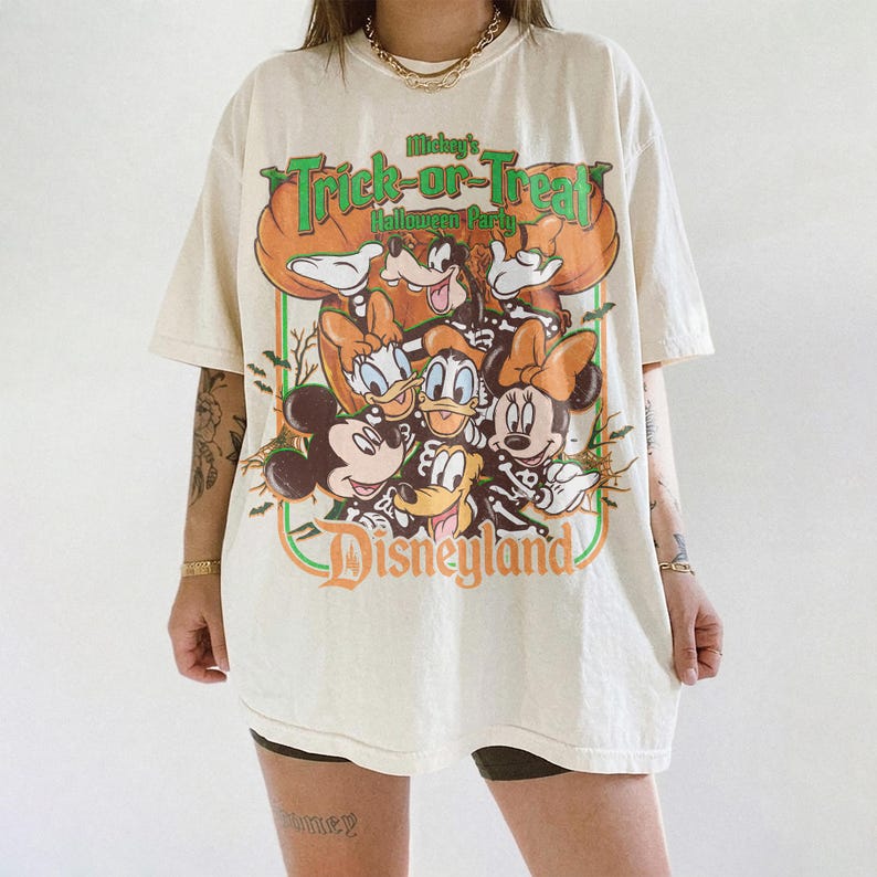 K&ouml;nnte beinhalten: Ein cremefarbenes Kurzarm-T-Shirt mit einem Grafikdruck von Mickey Maus, Minnie Maus, Donald Duck, Daisy Duck und Goofy in Skelettkost&uuml;men. Der Text lautet Mickey's Trick-or-Treat Halloween Party und Disneyland.
