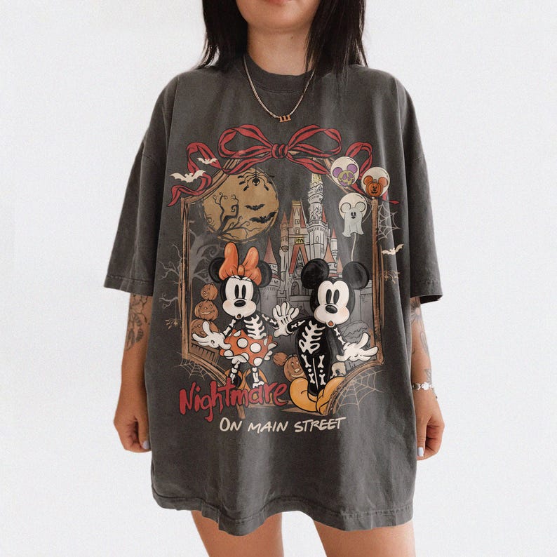 May include: Dark gray t-shirt featuring a Halloween-themed graphic. The design includes Mickey and Minnie Mouse dressed as skeletons, a haunted castle, and the words "Nightmare on Main Street."
