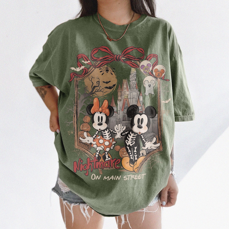 May include: Olive green t-shirt featuring a Halloween-themed graphic. The design includes Mickey and Minnie Mouse dressed as skeletons, a castle, pumpkins, and the text "Nightmare On Main Street". The shirt has short sleeves and a relaxed fit.