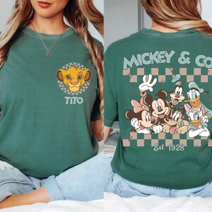 K&ouml;nnte beinhalten: Gr&uuml;nes T-Shirt mit einem Simba-Motiv und dem Namen "Tito". Auf der R&uuml;ckseite des Shirts befindet sich ein Mickey & Co.-Design mit Mickey, Minnie, Goofy, Donald und Daisy Duck. Der Text "Est. 1928" ist ebenfalls sichtbar.
