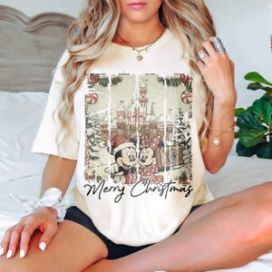 May include: Cream-colored t-shirt featuring a distressed graphic of Mickey and Minnie Mouse in front of a castle, with the words "Merry Christmas" below. The design includes Christmas trees and candy canes. A festive holiday tee.