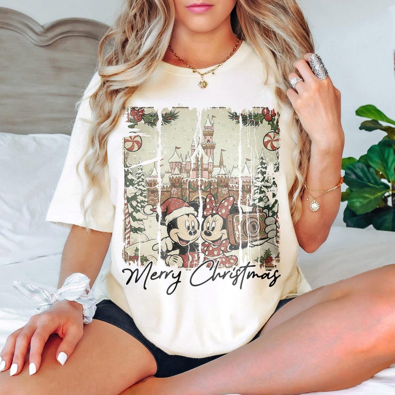 May include: Cream-colored t-shirt featuring a distressed graphic of Mickey and Minnie Mouse in front of a castle, with the words "Merry Christmas" below. The design includes Christmas trees and candy canes. A festive holiday tee.