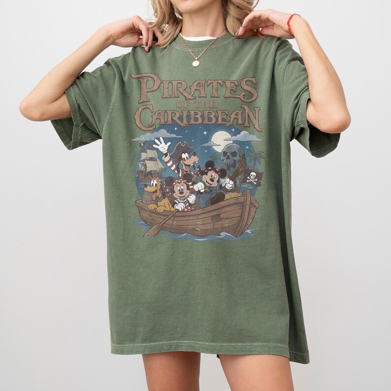 K&ouml;nnte beinhalten: Olivgr&uuml;nes T-Shirt mit einer Grafik von Disney-Figuren in einem Piratenschiff und dem Text "Pirates of the Caribbean". Das Design umfasst Micky Maus, Minnie Maus und Pluto. Das T-Shirt hat einen Vintage-Look.