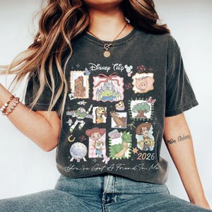 May include: Dark gray t-shirt featuring a collage of Disney characters and landmarks. The shirt includes the text "Disney Trip" and "2026", along with the phrase "You've Got A Friend In Me".