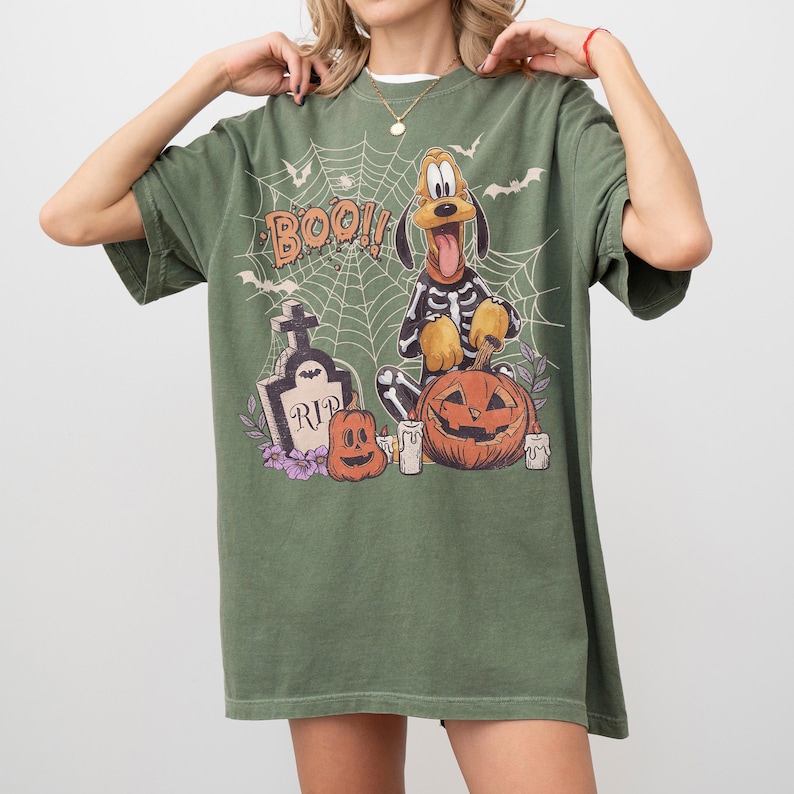 May include: An olive green t-shirt featuring a graphic print of a cartoon dog wearing a skeleton costume, a spider web, bats, a tombstone with the text RIP, a carved pumpkin, and the word BOO. The shirt has short sleeves and a crew neckline.