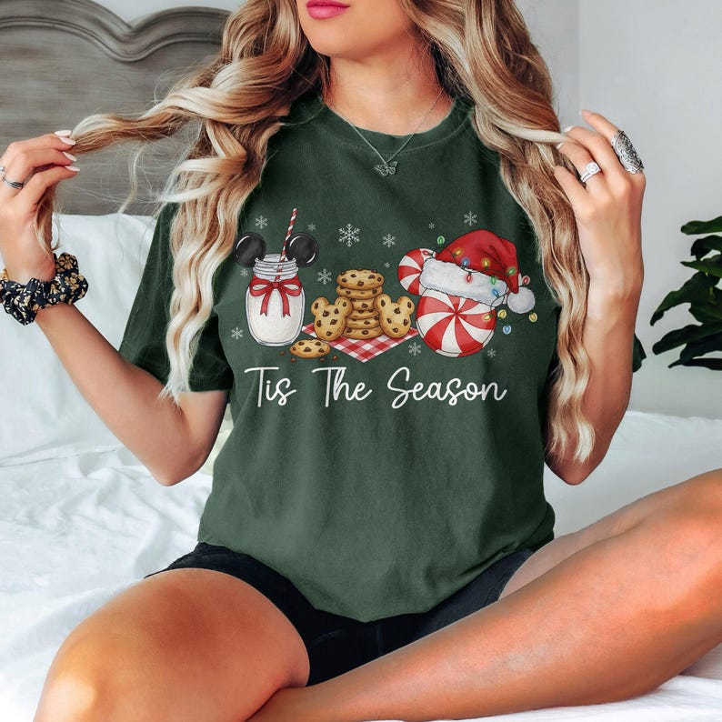 May include: Dark green t-shirt with a festive graphic. The design features a jar of milk with Mickey Mouse ears, cookies, and a Santa Claus-themed candy cane. The text "Tis The Season" is written in white script.
