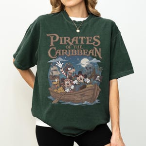 K&ouml;nnte beinhalten: Dunkelgr&uuml;nes T-Shirt mit dem Text "Pirates of the Caribbean" &uuml;ber einer Cartoon-Illustration von Figuren in einem Boot. Die Illustration zeigt einen Totenkopf, ein Piratenschiff und einen Mond.