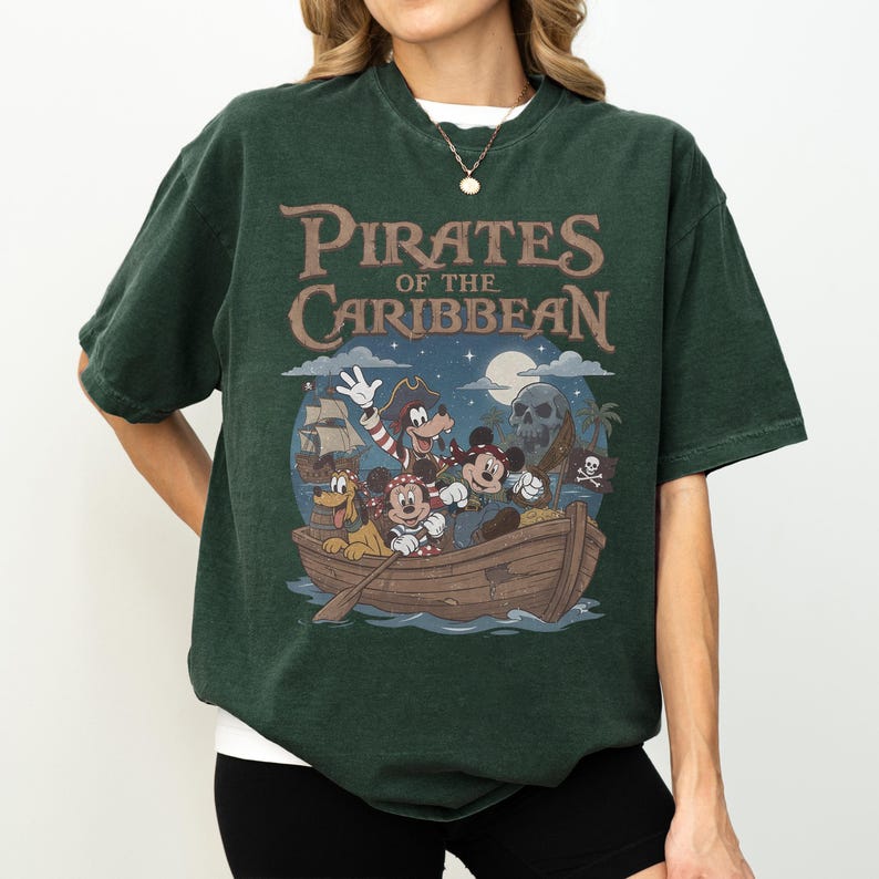 K&ouml;nnte beinhalten: Dunkelgr&uuml;nes T-Shirt mit dem Text "Pirates of the Caribbean" &uuml;ber einer Cartoon-Illustration von Figuren in einem Boot. Die Illustration zeigt einen Totenkopf, ein Piratenschiff und einen Mond.