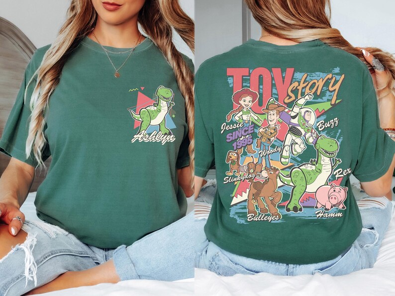 K&ouml;nnte beinhalten: Gr&uuml;nes T-Shirt mit einem Cartoon-Dinosaurier und dem Namen "Ashlyn" auf der Vorderseite. Auf der R&uuml;ckseite sind das "Toy Story"-Logo und Charaktere wie Woody, Buzz und Rex abgebildet. Das Shirt ist ein bequemer, l&auml;ssiger Stil.