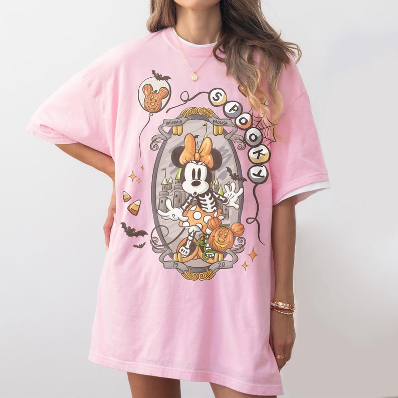 May include: Pink t-shirt featuring a Halloween-themed Minnie Mouse graphic. The design includes a skeleton Minnie, a pumpkin, and the words "Spotlight". The shirt has a double-layered look with white sleeves.