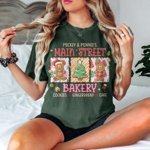 Op de afbeelding: Donkergroen T-shirt met de tekst "Mickey & Minnie's Main Street Bakery". Het shirt heeft een afbeelding van peperkoekpersonages en een kerstboom. De woorden "Cookies, Gingerbread, Cake" staan eronder gedrukt.