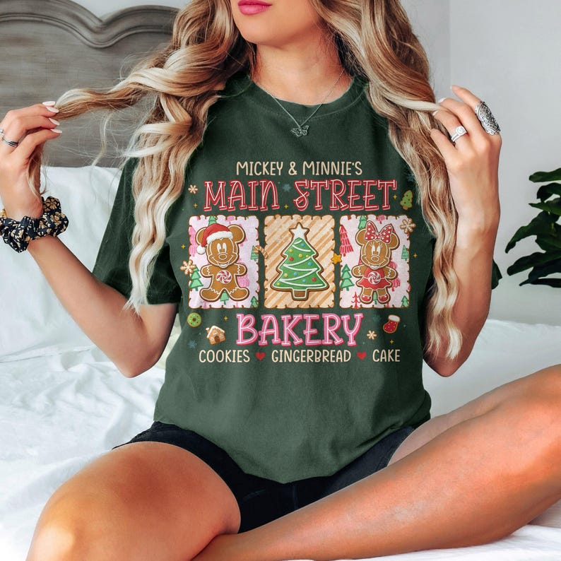 Op de afbeelding: Donkergroen T-shirt met de tekst "Mickey & Minnie's Main Street Bakery". Het shirt heeft een afbeelding van peperkoekpersonages en een kerstboom. De woorden "Cookies, Gingerbread, Cake" staan eronder gedrukt.