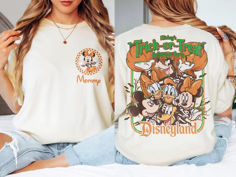 May include: A cream-colored t-shirt featuring a small circular graphic on the front left chest with Minnie Mouse and the word Mommy. The back displays a large graphic of Mickey, Minnie, Donald, Daisy, and Goofy in skeleton costumes with the text Mickey's Trick-or-Treat Halloween Party Disneyland.