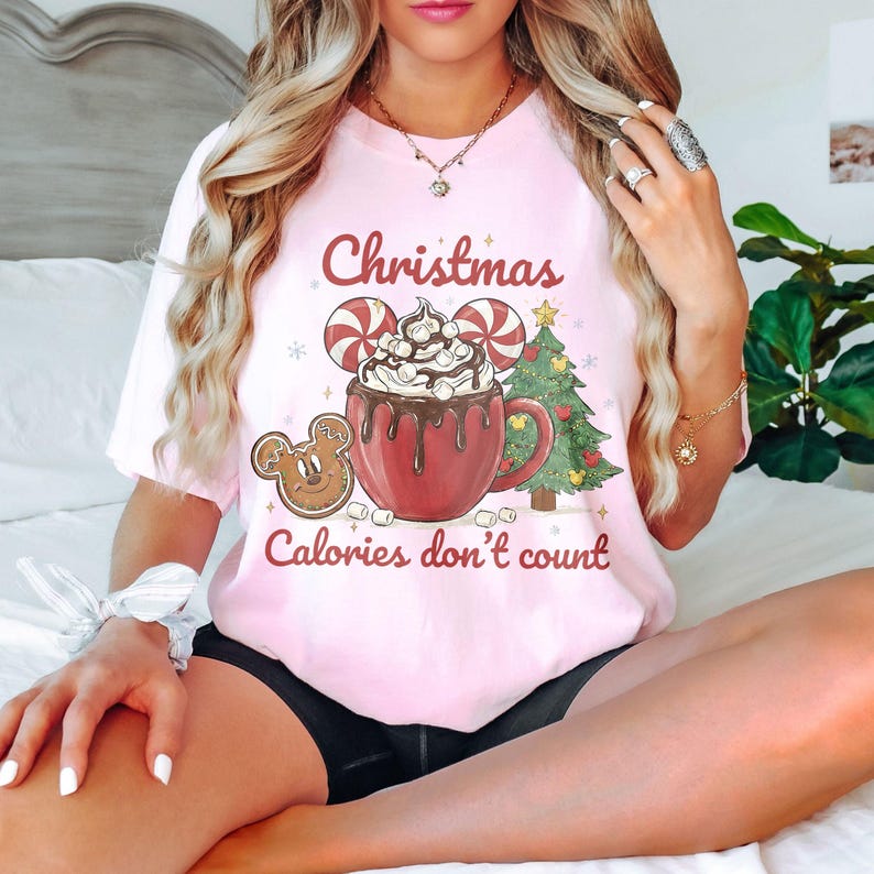 May include: Pink t-shirt with a Christmas-themed graphic. The design includes a red mug of hot chocolate topped with whipped cream, candy canes, a gingerbread cookie, and a Christmas tree. Text reads "Christmas Calories don't count."